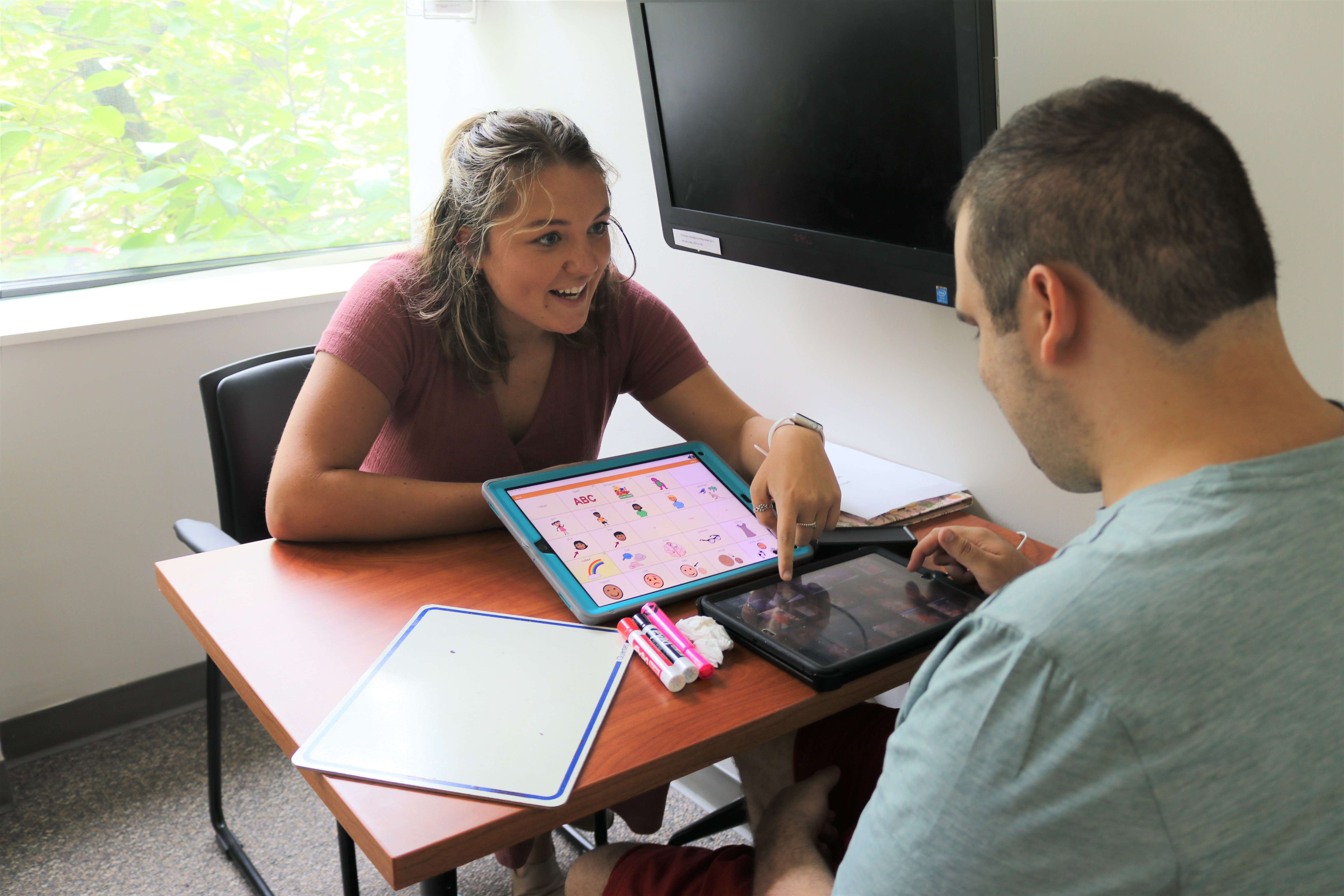 SLP sitting with client Daniel instructing him how to use his new AAC device to communicate